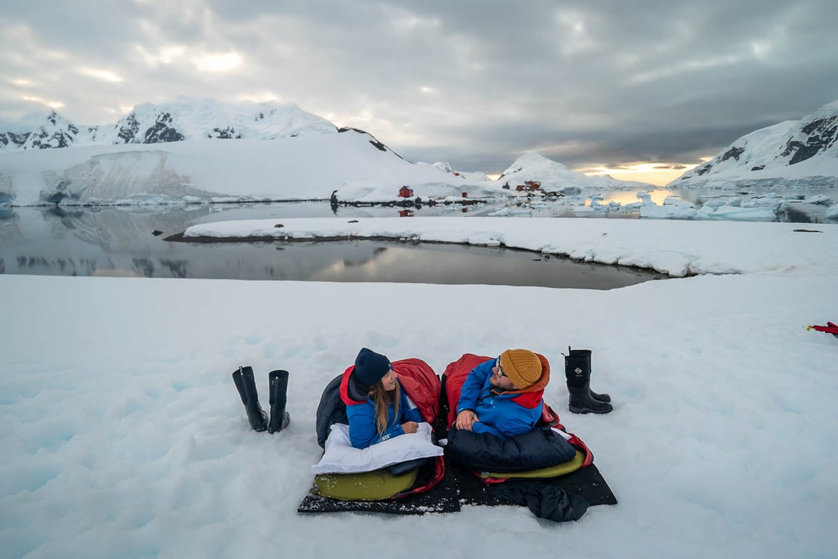 Couple laying in sleeping bags with a research station in the background in Paradise Bay, Antarctica