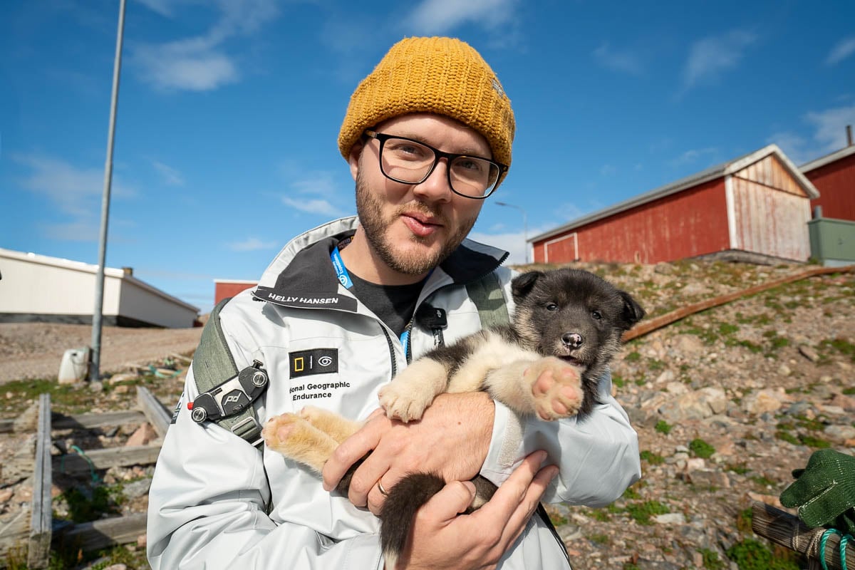 Man holding a sled dog puppy with colorful buildings in the background in Ittoqqortoormiit, Greenland