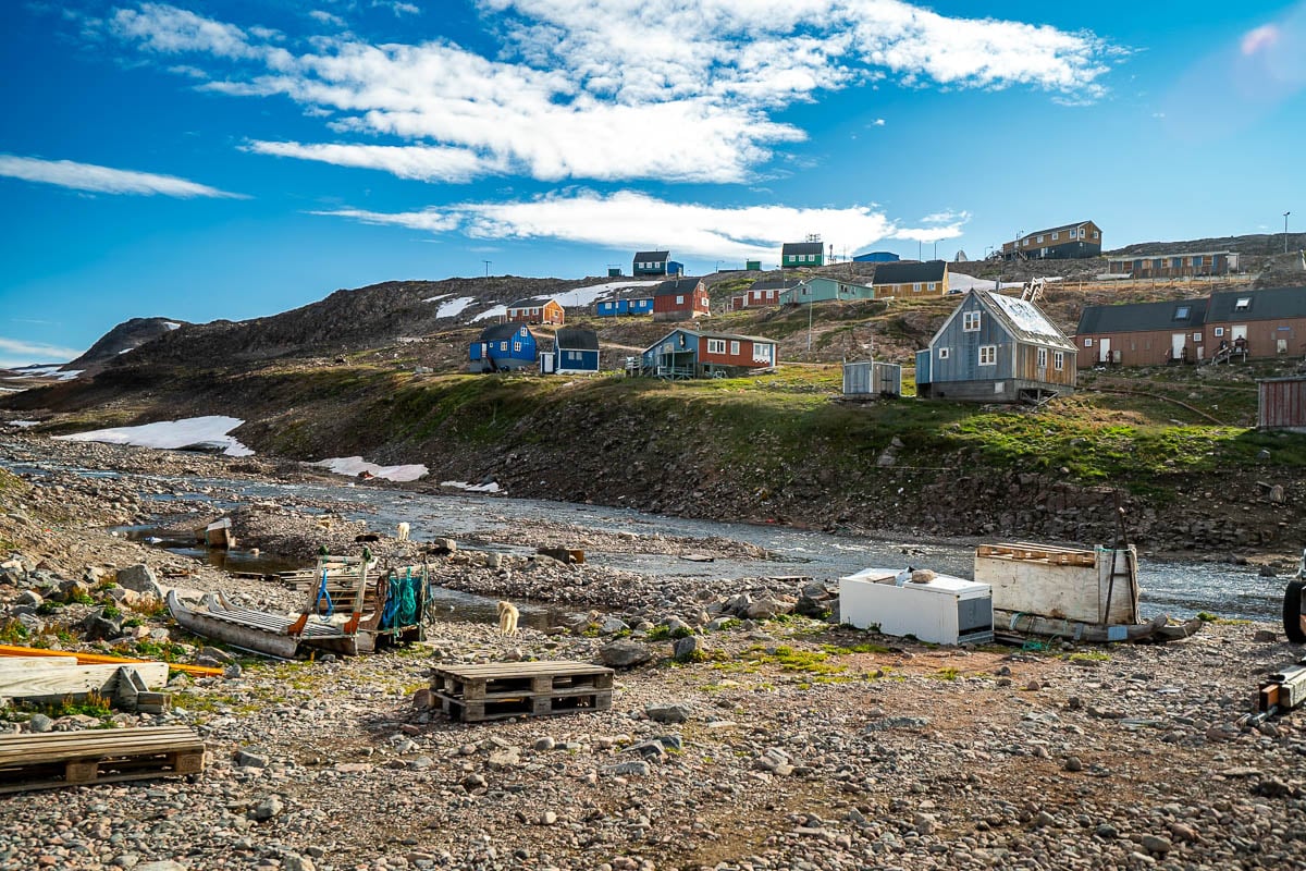 Dog sleds and dogs standing next to a stream with colorful buildings in the background in Ittoqqortoormiit, Greenland