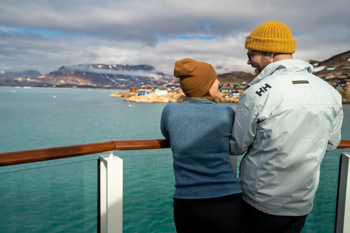 Couple standing on a cruise ship with colorful buildings on a Rocky Mountain slope in Ittoqqortoormiit, Greenland
