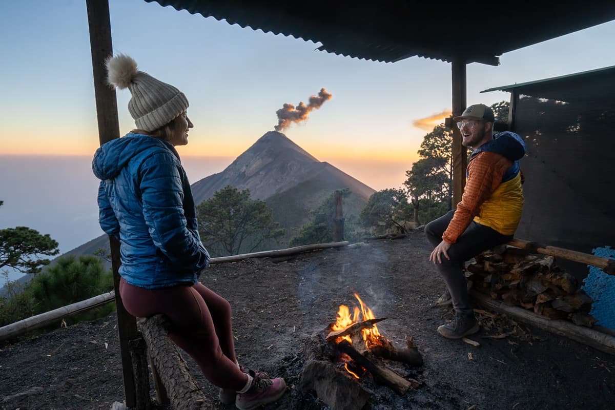 Couple hanging around a campfire while the Fuego Volcano erups in the background along the Acatenango hike in Guatemala