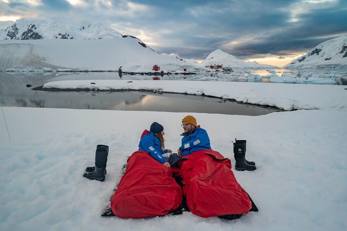Couple laying in bivy bags in the snow with snowy mountains in the background on a camping trip on an Antarctica cruise