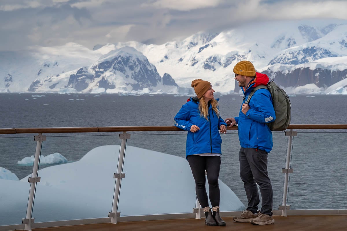 Couple standing on the top deck of a ship with icebergs and mountains in the background on an Antarctica cruise