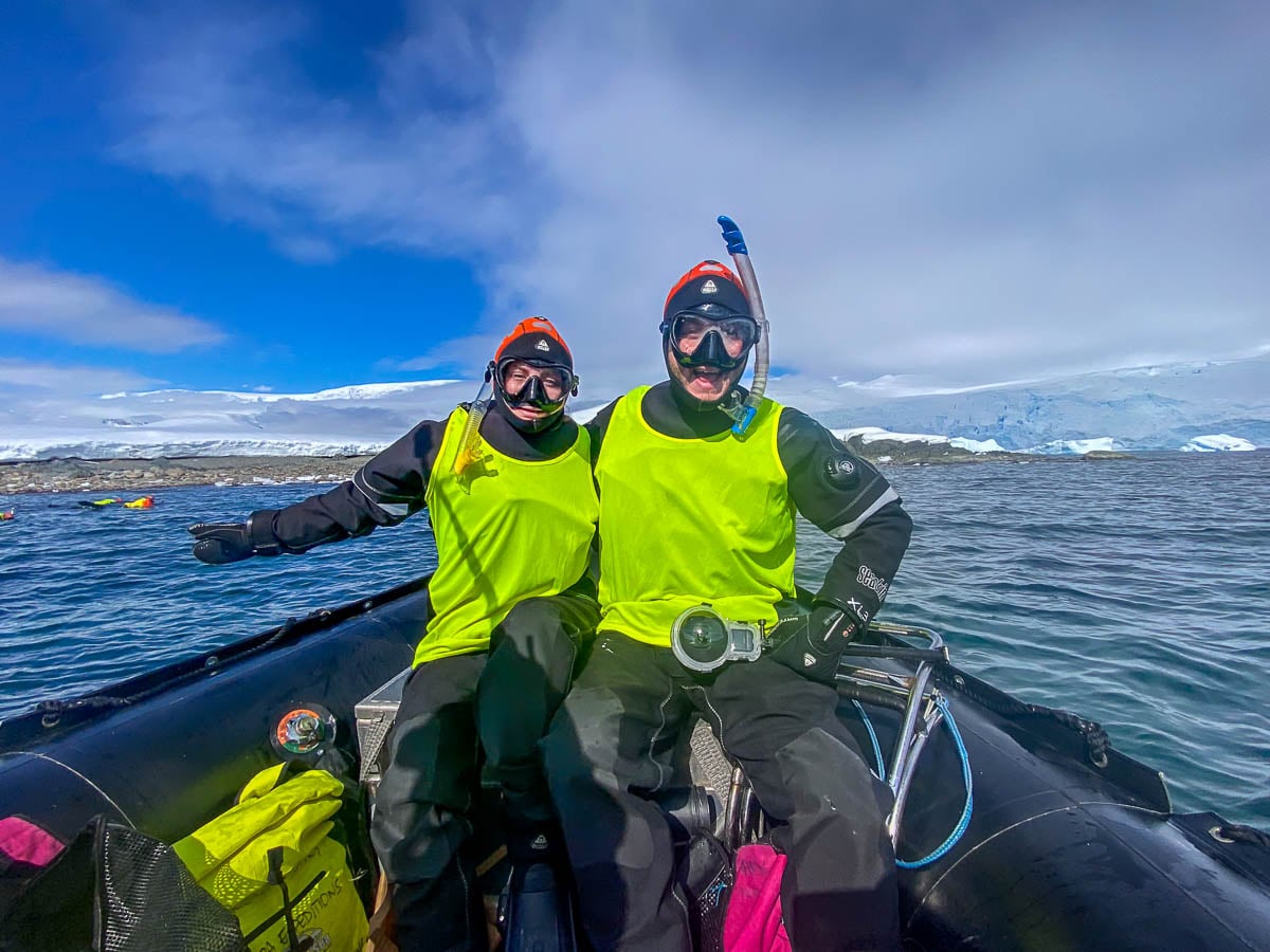 Couple wearing dry suits on a Zodiac ship with snowy mountains in the background