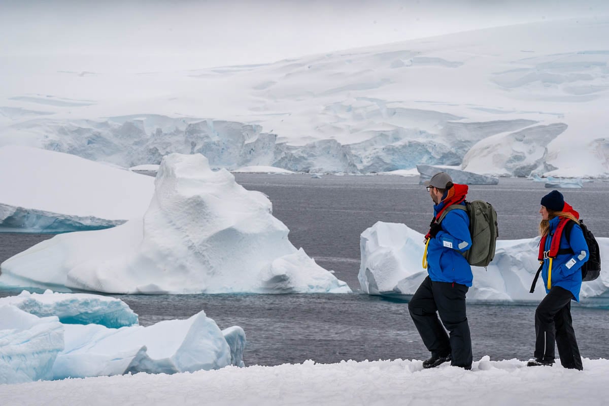 Couple walking on a snowy hill with icebergs and glaciers in the background in Antarctica