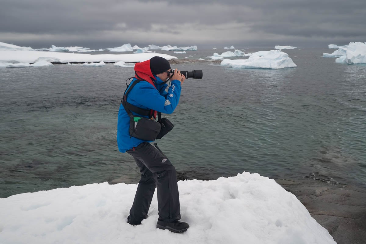 Person taking a photo on a snowy outcropping with icebergs in the background in Antarctica
