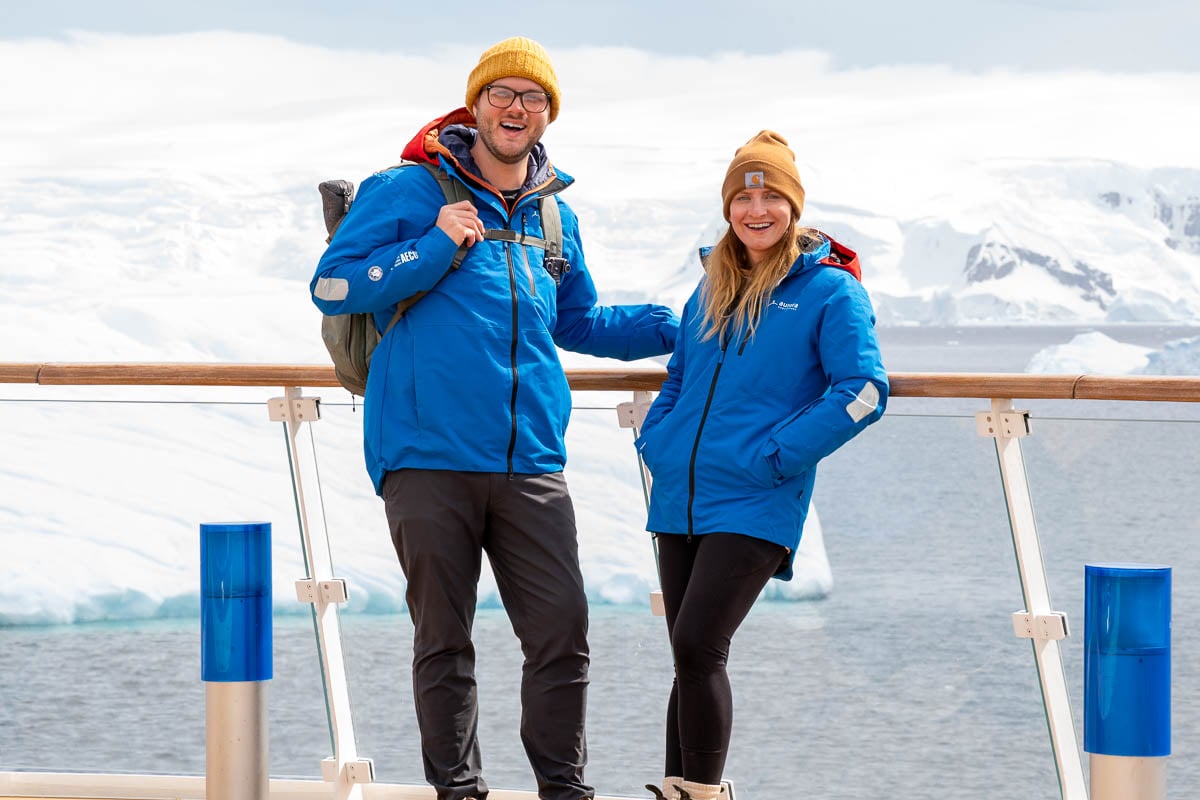 Couple standing on the top deck of the Aurora Expeditions Greg Mortimer ship with icebergs in the background in Antarctica