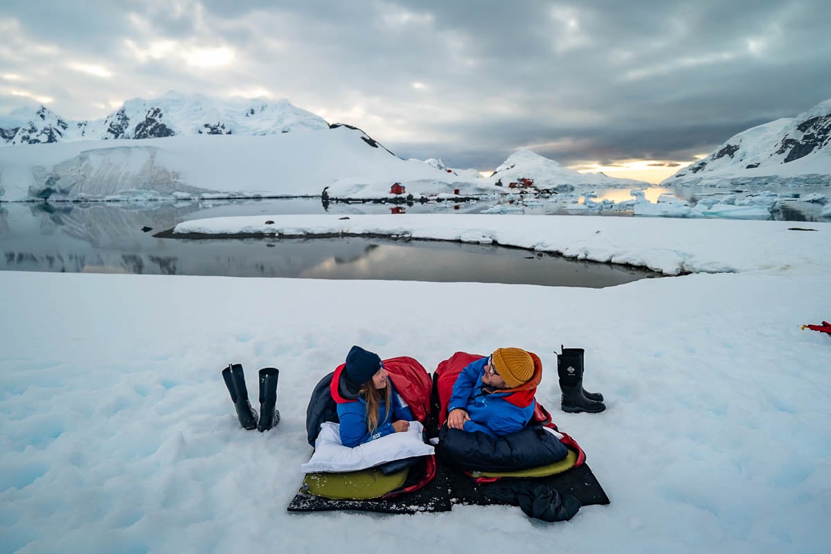 Couple laying down in sleeping bags on Paradise Bay with snowy mountains in the background in Antarctica