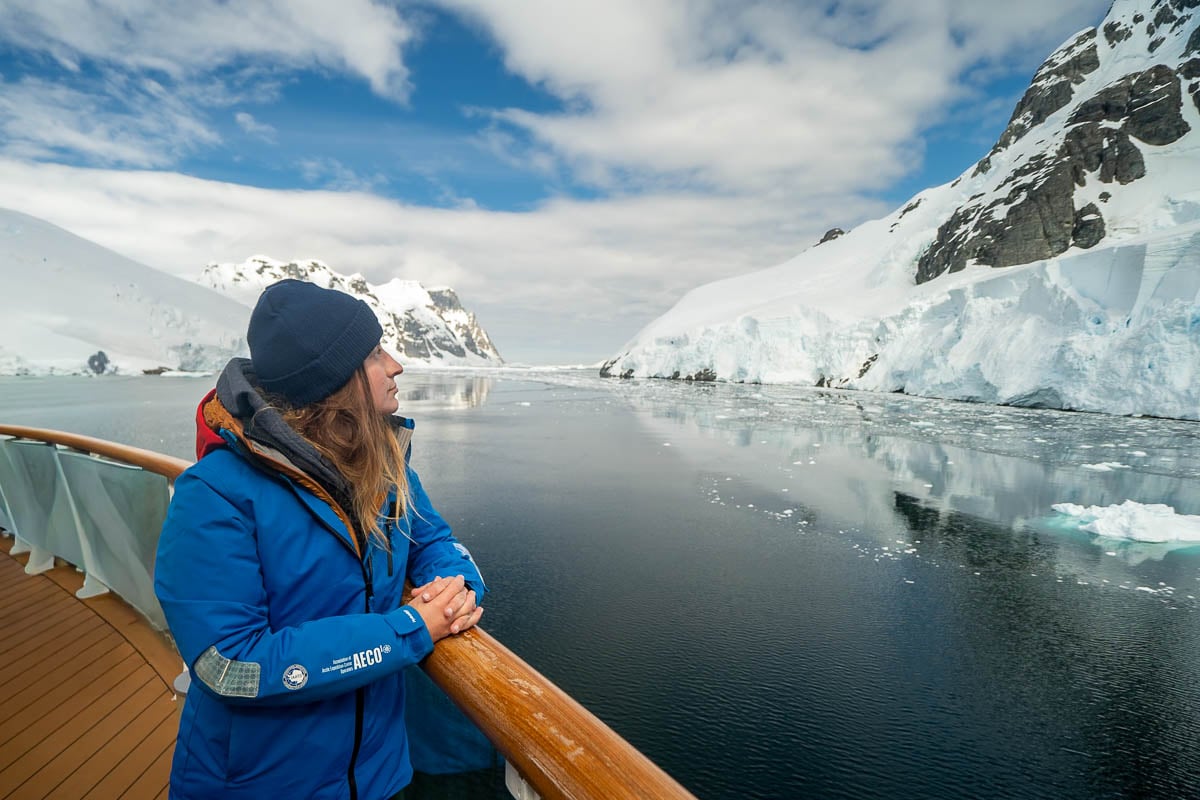 Woman standing next to the railing on the top deck of the Aurora Expeditions Greg Mortimer ship while looking at glaciers in the Lemaire Channel in Antarctica