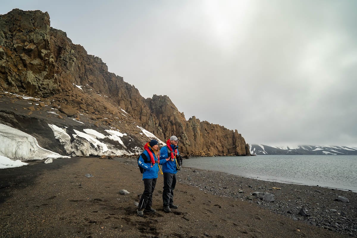Couple standing on a volcanic beach with rocky cliffside at Deception Island on an Aurora Expeditions Antarctica cruise
