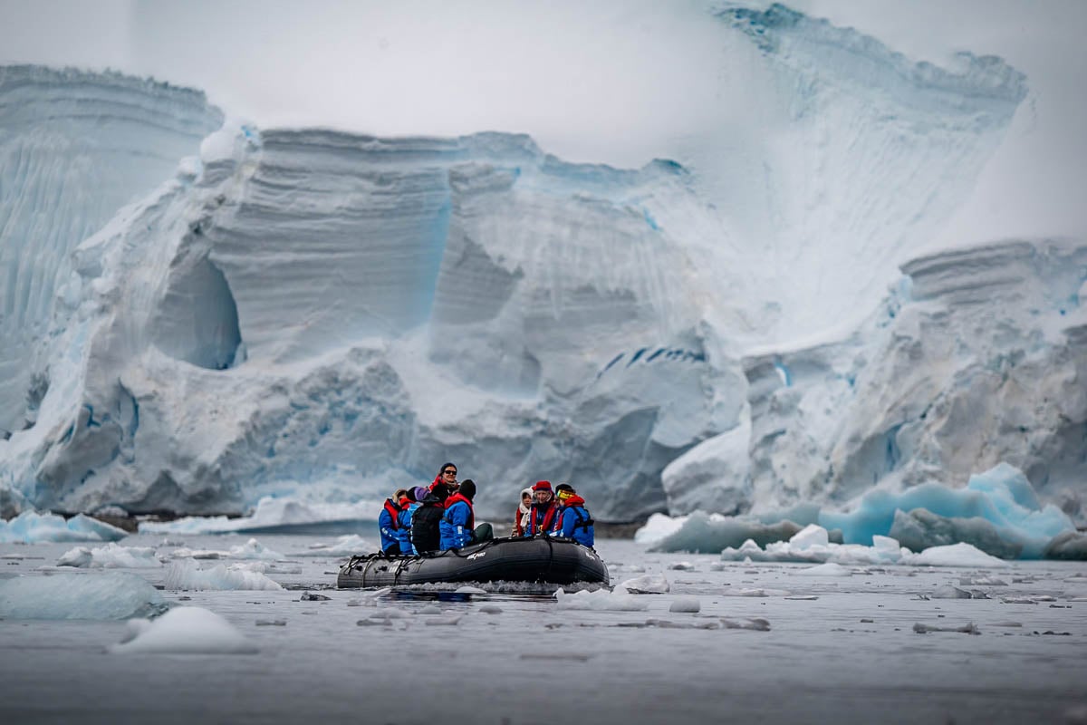 Expedition guide with passengers on Aurora Expeditions with rocky cliffs and icebergs in the background in 5. Chiriguano Bay, Antarctica