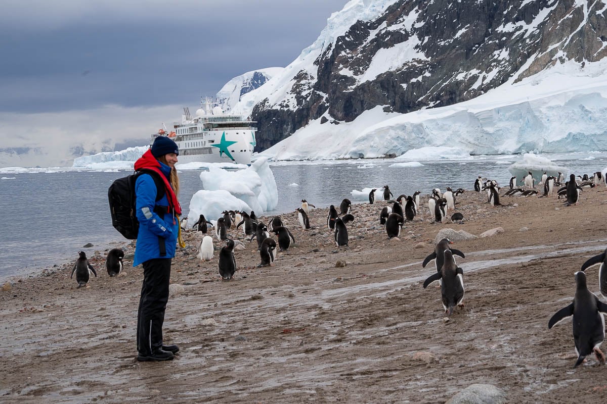 Woman smiling while watching gentoo penguins waddle with the Aurora Expeditions Greg Mortimer ship and a snowy mountain in the background at Neko Harbor, Antarctica