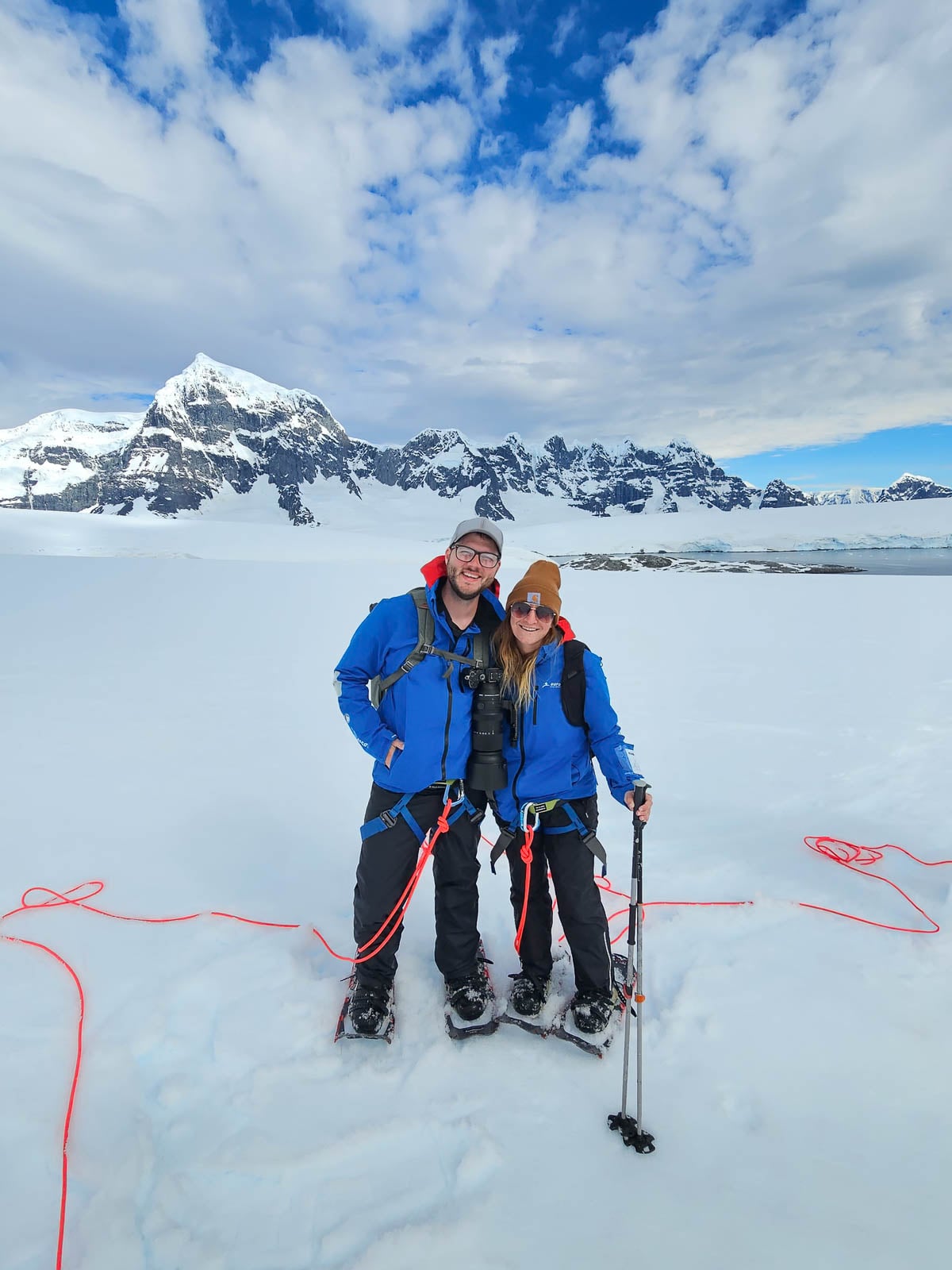 Smiling couple wearing snowshoes and harnessed with snowy mountains in the background at Damoy Point. Antarctica