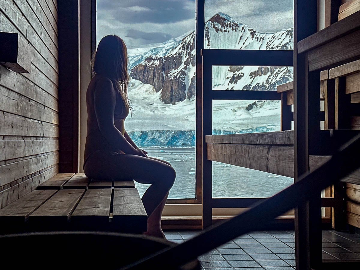 Woman sitting on a wooden bench in a sauna with a snowy mountain out of the window on the Aurora Expeditions Greg Mortimer ship in Antarctica