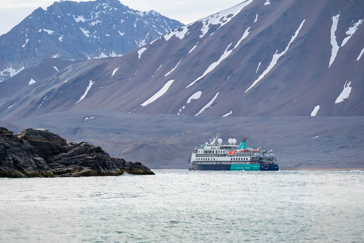 Aurora Expeditions Sylvia Earle ship with a volcanic cliffside in the foreground and snowy mountains in the background in Svalbard, Norway