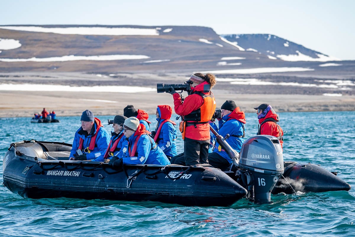 Aurora Expeditions guide taking photos with a camera while guests sit on a Zodiac with mountains in the background in Svalbard, Norway