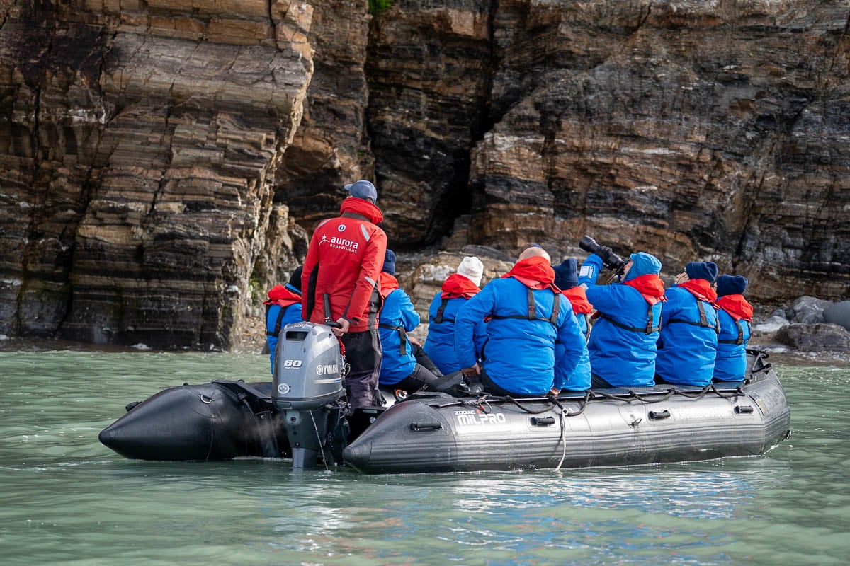 Aurora Expeditions guide with guests sitting on a Zodiac boat with a rugged cliffside in the background in Svalbard, Norway