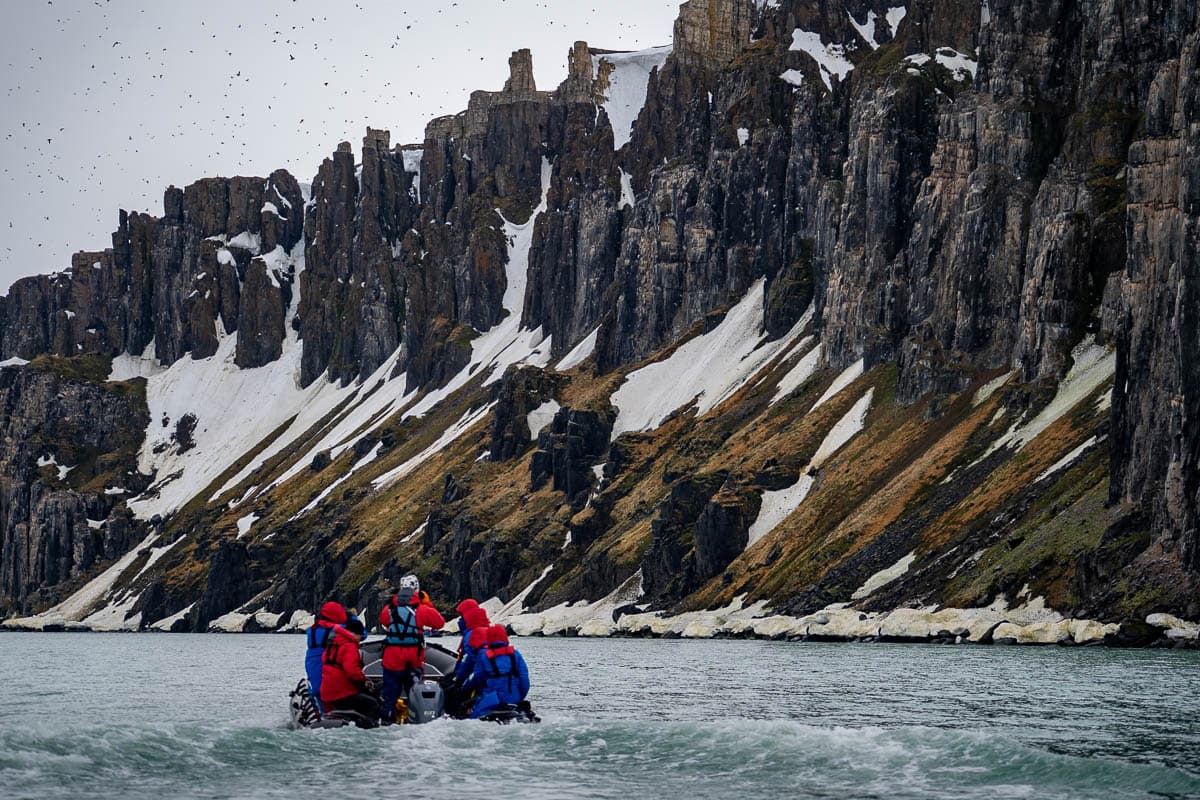 Zodiac cruising towards volcanic cliffs covered with patches of snow at Alkefjellet with birds flying around in Svalbard, Norway