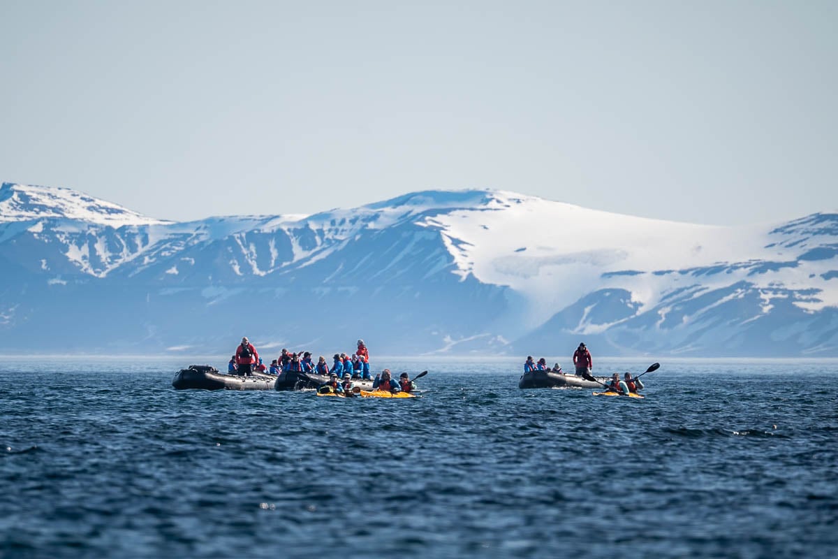 Kayakers and Zodiac boats with Aurora Expeditions guides cruise in front of snowy mountains in the background in Svalbard, Norway