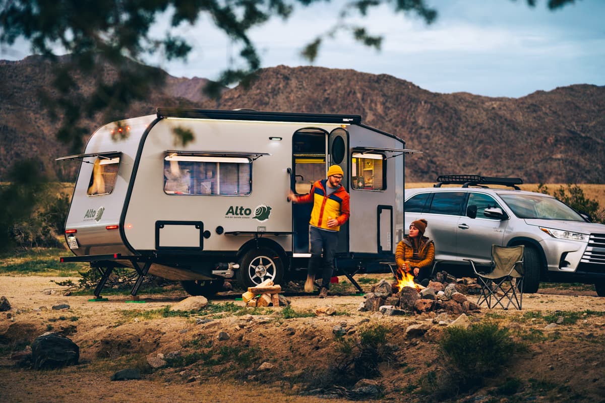 Man stepping out of a Safari Condo Alto A1852 with a woman sitting around a campfire with mountains in the background in Joshua Tree, California