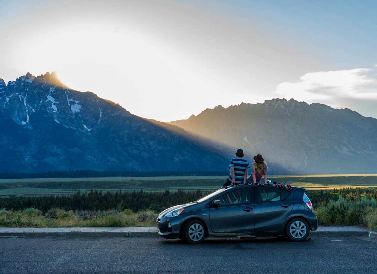 Couple sitting on top of a Toyota Prius with the sun setting over the Teton Mountains in Grand Teton National Park in Wyoming