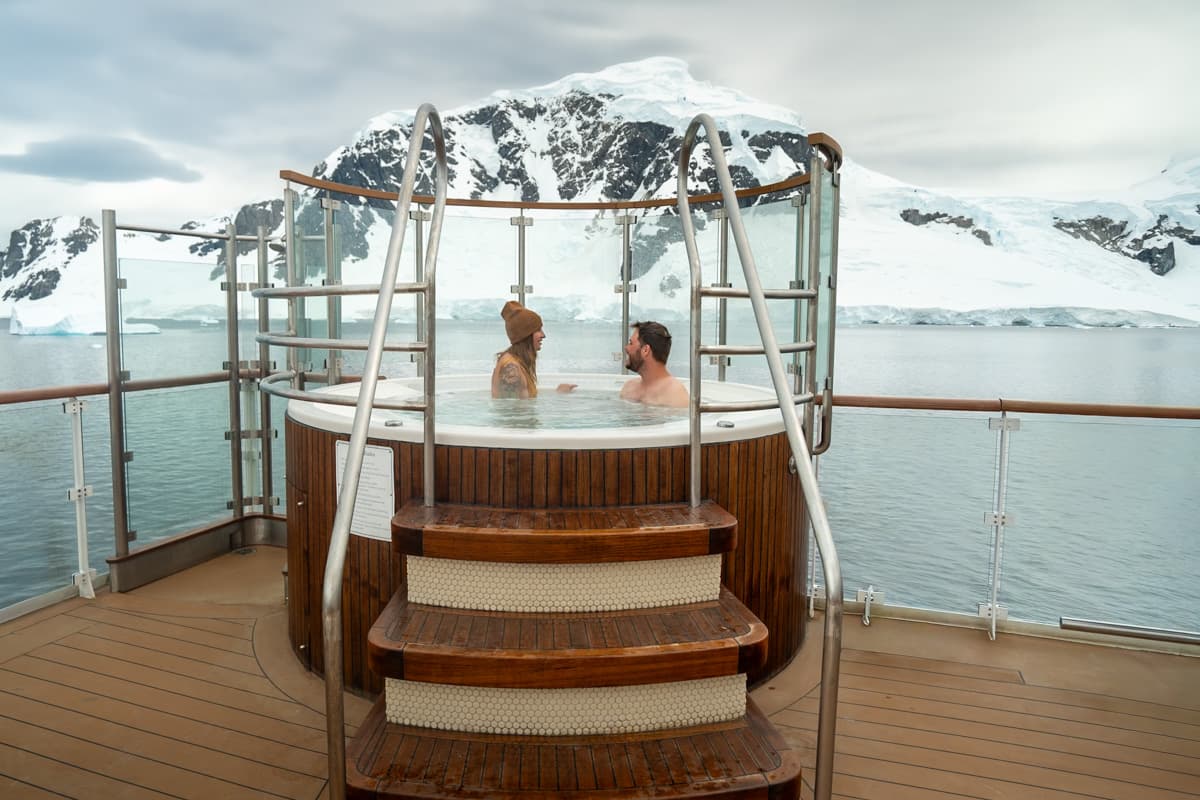 Couple sitting in a hot tub on the top deck of a ship with a snowy mountain in the background in Antarctica
