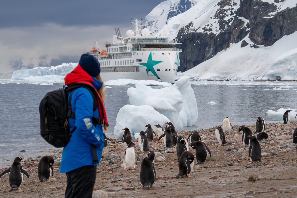 Woman standing on a rocky beach with gentoo penguins and an Aurora Expeditions ship in the background in Neko Harbor, Antarctica