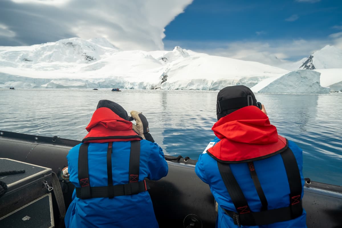 Passengers kneeling in a Zodiac boat taking photos of a whale with snow covered mountains in the background in Antarctica