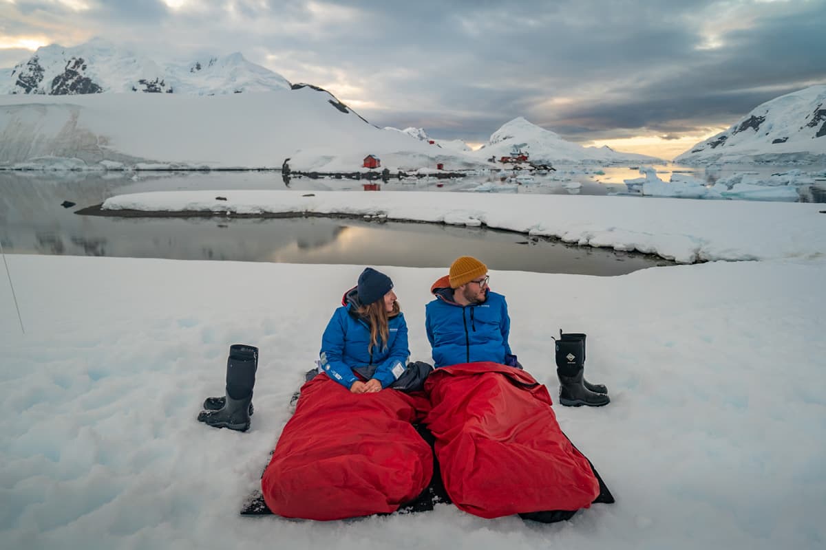 Couple sitting in sleeping bags on top of snow with snowy mountains in the background in Paradise Bay, Antarctica