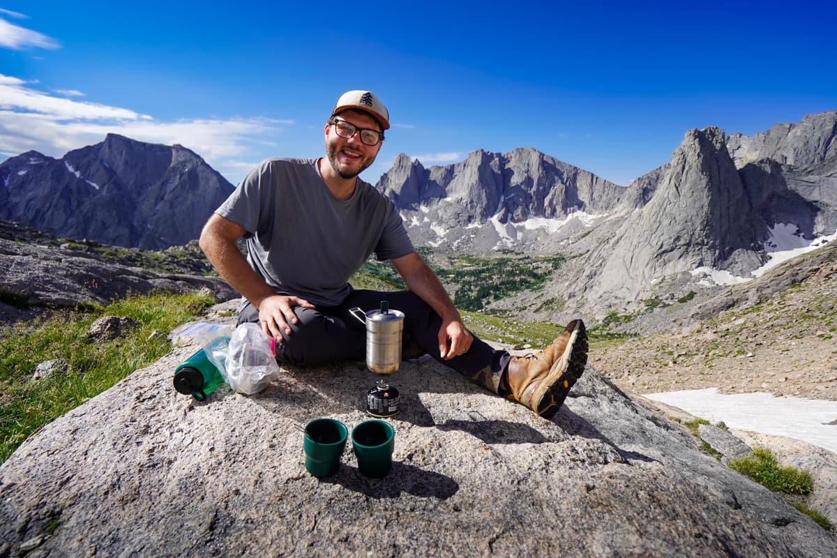 Man sitting on a rocky outcropping with campstove and coffee setup with the Cirque of the Towers in the background in the Wind River Range in Wyoming