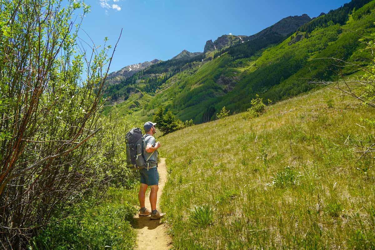Man with a backpacking back hiking along the Conundrum Hot Springs trail with snowcapped mountains in the background in Maroon Bells-Snowmass Wilderness in Aspen, Colorado