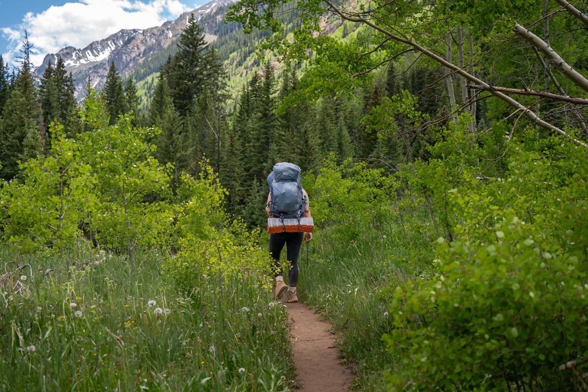 Woman with a backpacking back hiking along the Conundrum Hot Springs trail with snowcapped mountains in the background in Maroon Bells-Snowmass Wilderness in Aspen, Colorado