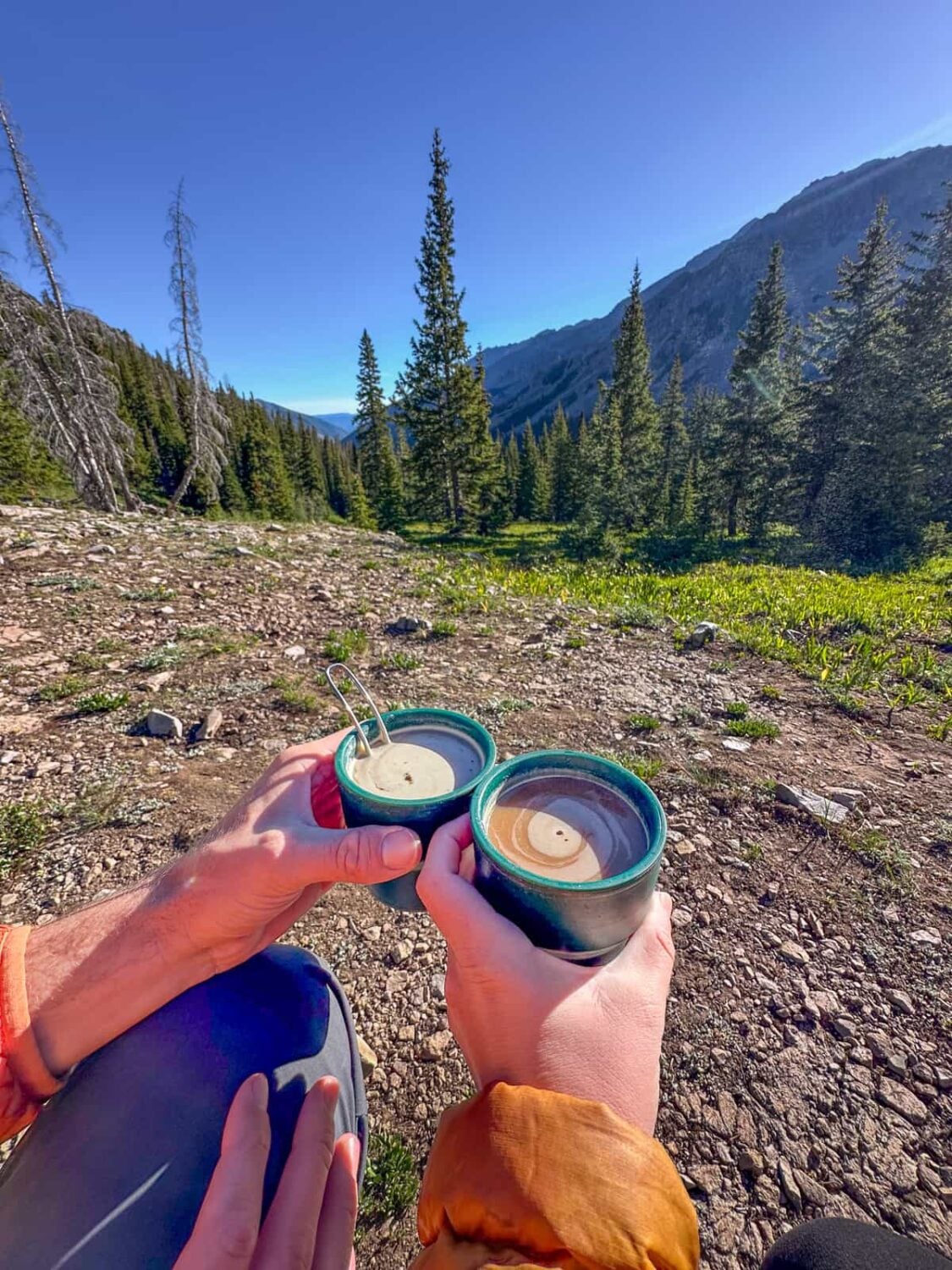 Couple holding coffee cups at a campsite at Conundrum Hot Springs Trail with mountains in the background in Maroon Bell-Snowmass Wilderness in Aspen, Colorado