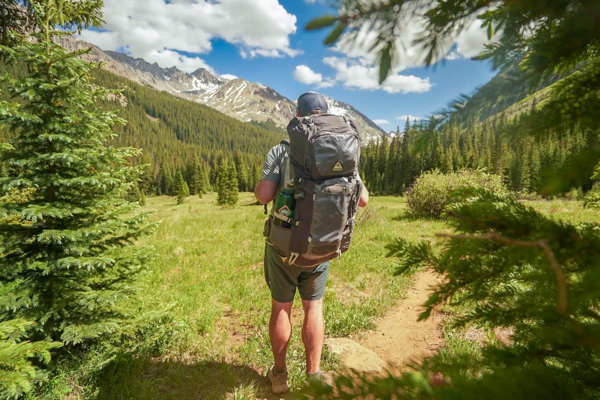 Man with a backpack standing on the Conundrum Hot Springs Trail with snowcapped mountains in the background in Maroon Bells-Snowmass Wilderness in Aspen, Colorado