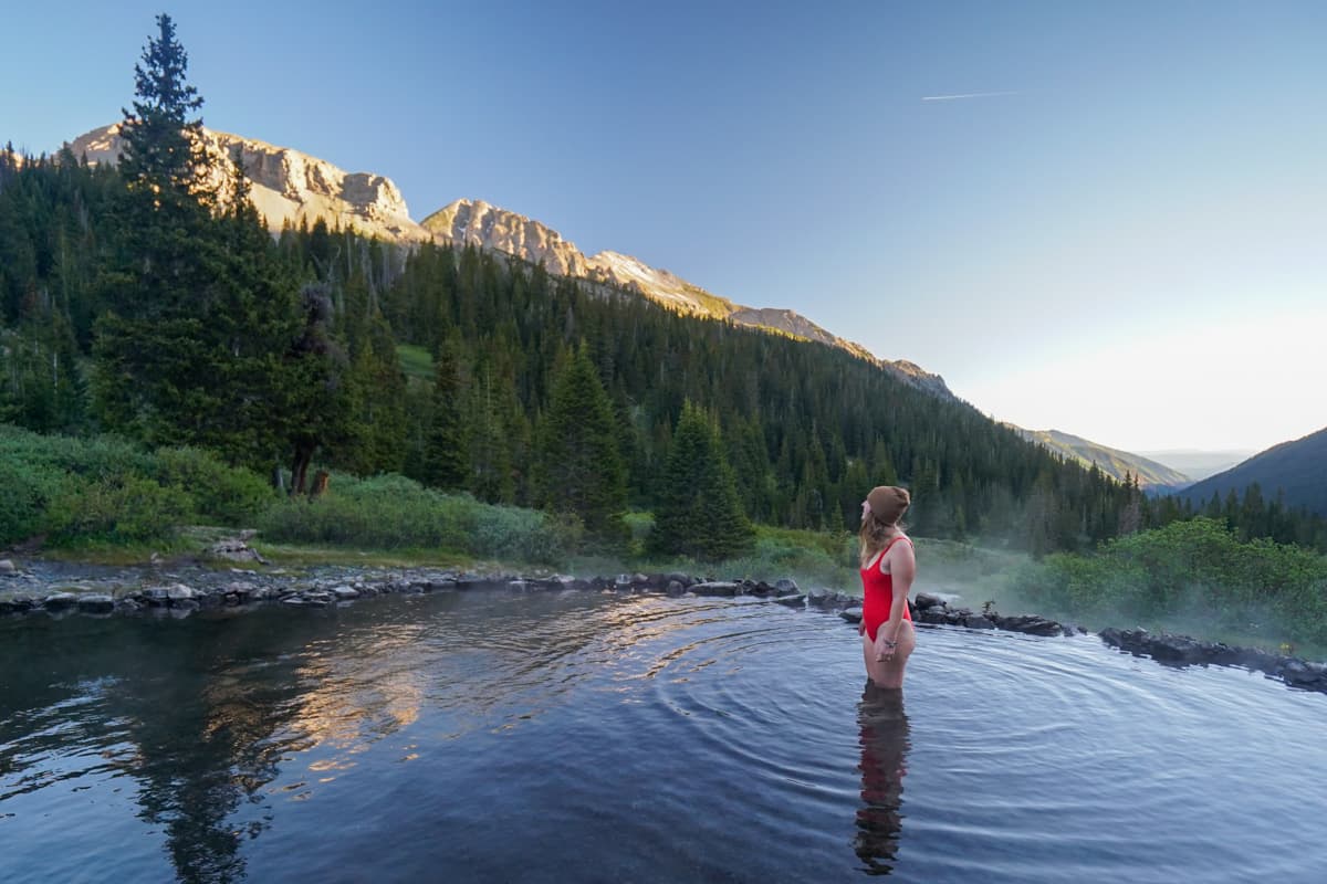 Woman standing in Conundrum Hot Springs with mountains with alpine glow on them in Maroon Bells-Snowmass Wilderness in Aspen, Colorado