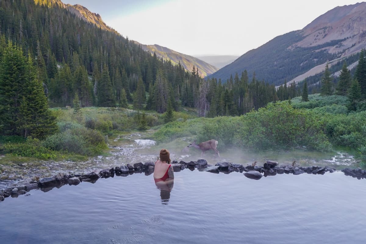 Woman soaking in Conundrum Hot Springs while a deer drinks from a stream nearby with mountains in the background in Maroon Bells-Snowmass Wilderness in Aspen, Colorado