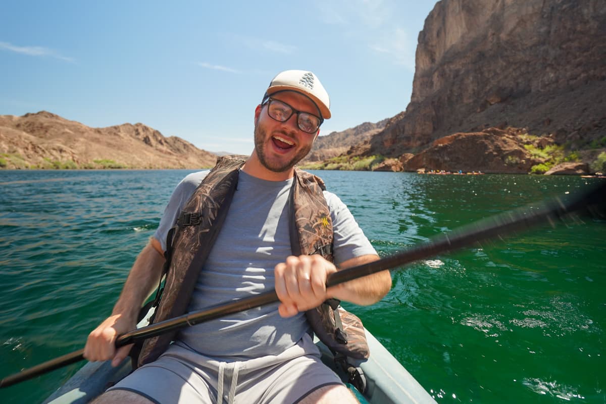 Smiling man sitting in a kayak paddling down the Colorado River with red cliff formations in the background in the Lake Mead National Recreation Area in Willow, Arizona