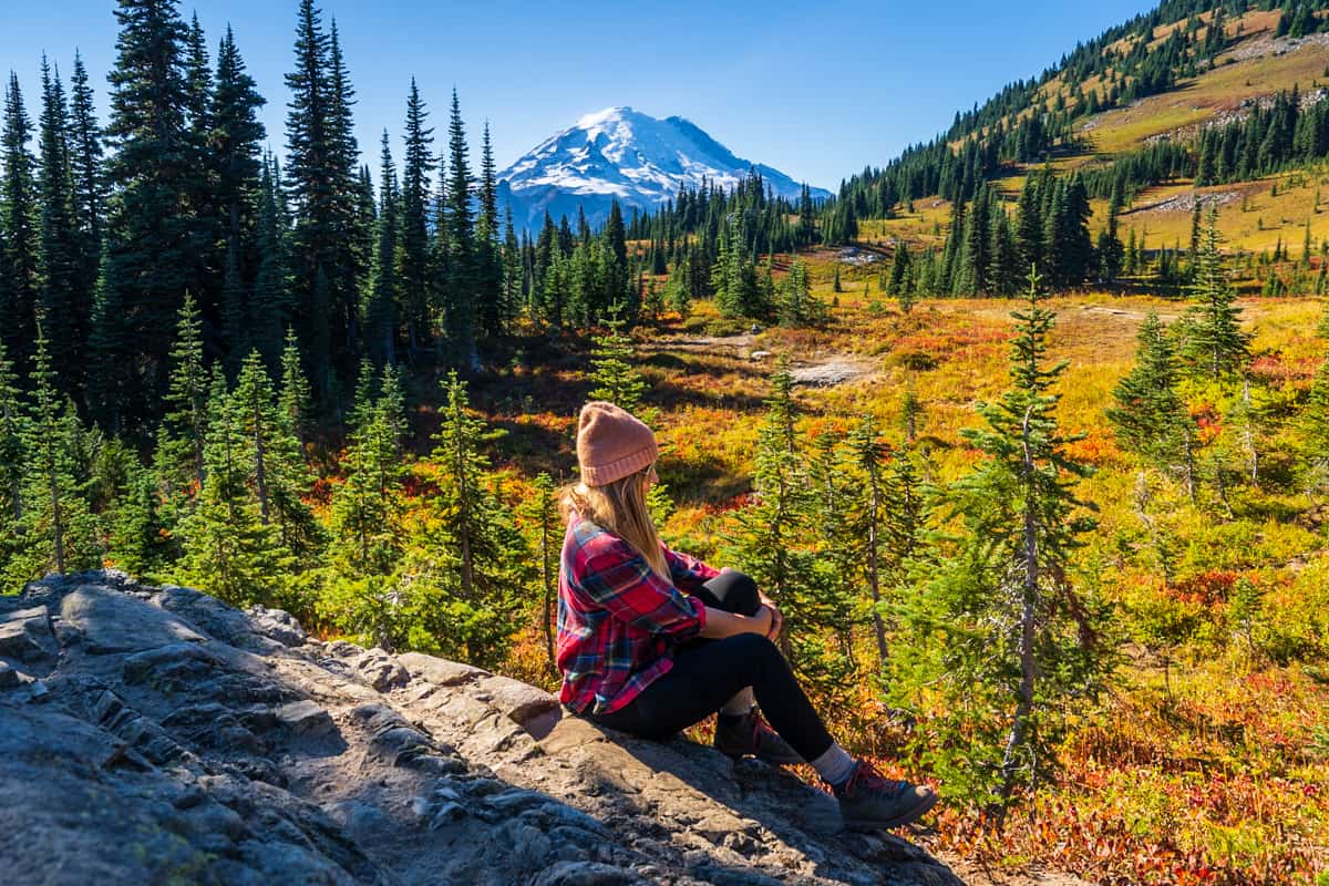 Woma nsitting on a rocky outcropping along the Naches Peak Loop Trail with Mount Rainier in the background in fall in Mount rainier National Park in Washington