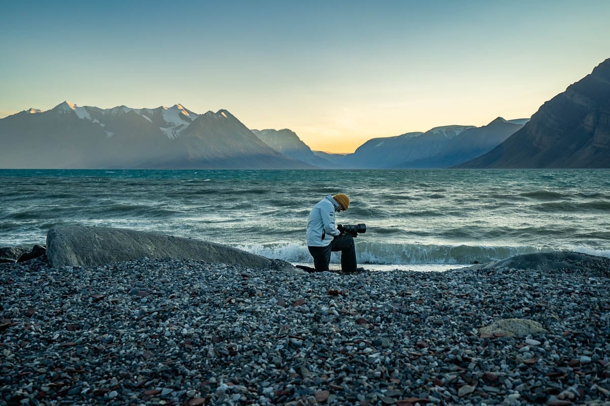 Man with a camera kneeling on a rocky beach with mountains in the background at sunset in Northeast Greenland National Park in Greenland