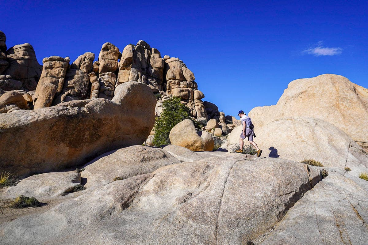 Man walking up a boulder with a rock formation in the background along the Hidden Valley Trail in Joshua Tree National Park, California