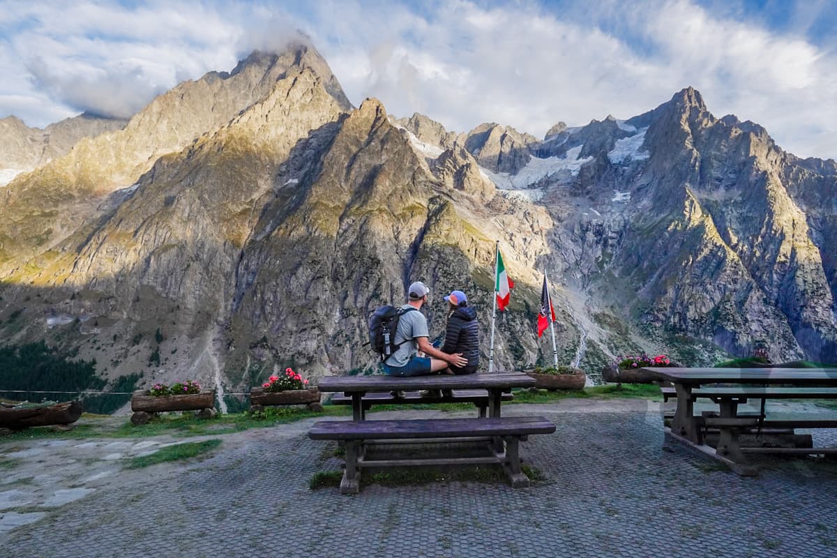 Couple sitting on a picnic table at Rifugio Bonatti, Italy with the Mont Blanc massif in the background along the Tour du Mont Blanc