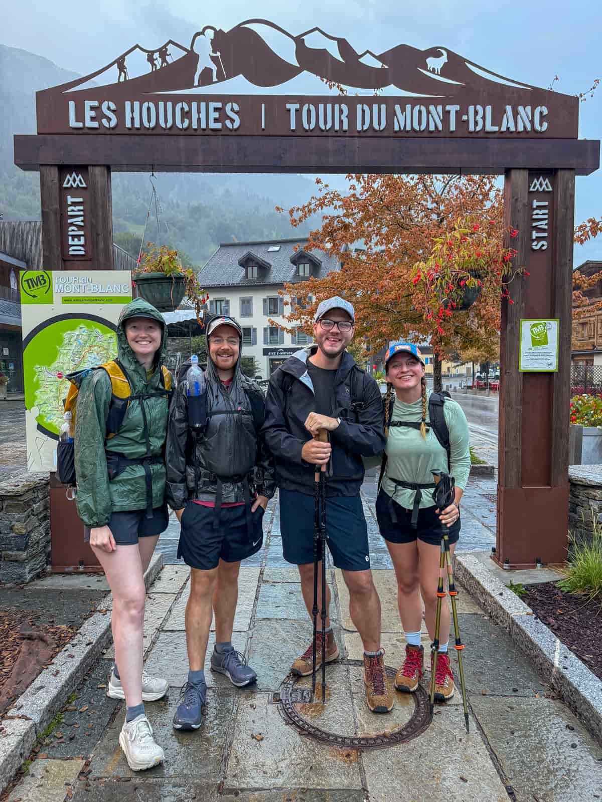 Four smiling friends near the Tour du Mont Blanc starting arch in Les Houches, France on a rainy day