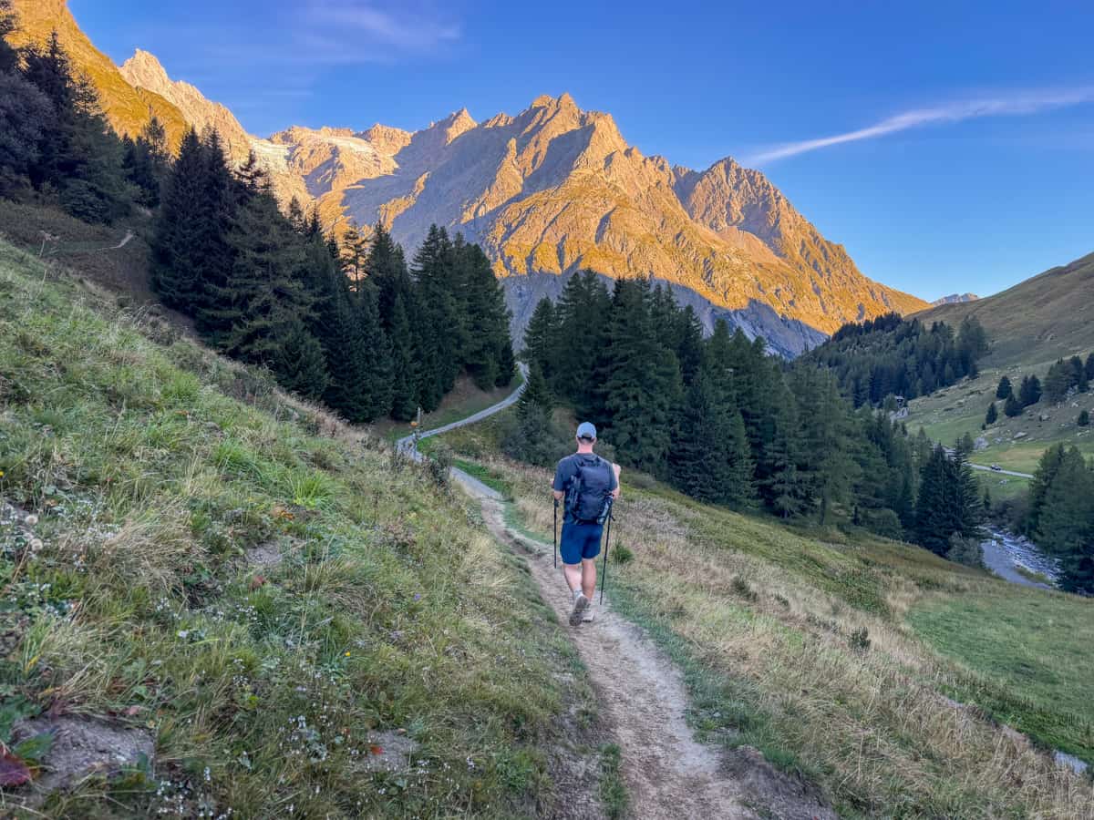 Man hiking on a dirt path with Swiss Alps in the background, illuminated by the rising sun along the Tour du Mont Blanc