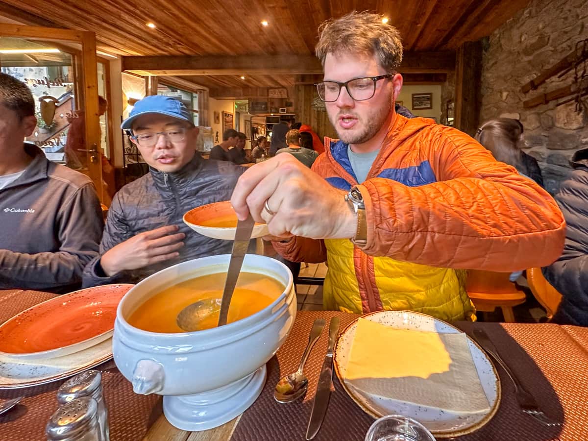 Man ladling soup into a bowl at a table at the Auberge de la Nova in Les Chapieux, France along the Tour du Mont Blanc