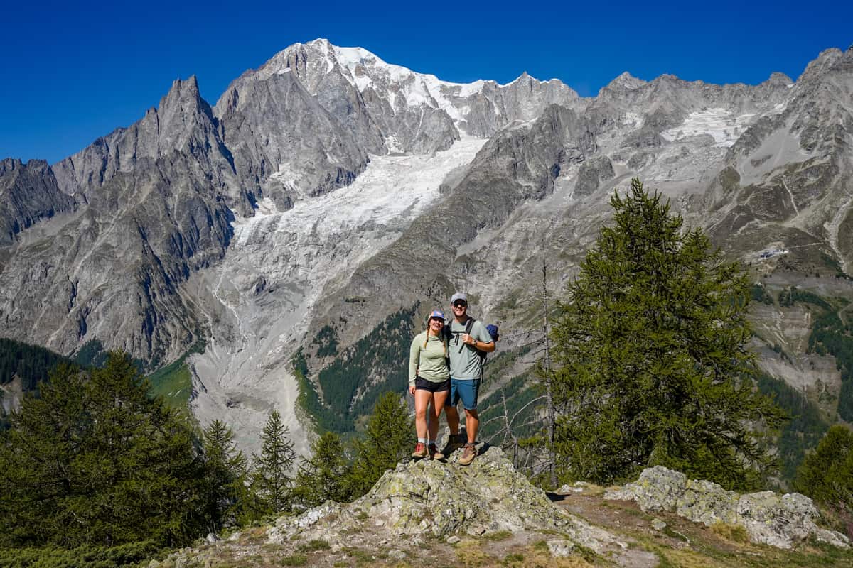 Smiling couple standing on a rocky outcropping with a large glacier and rugged mountains in the background in the Val Ferret above Courmayeur, Italy along the Tour du Mont Blanc