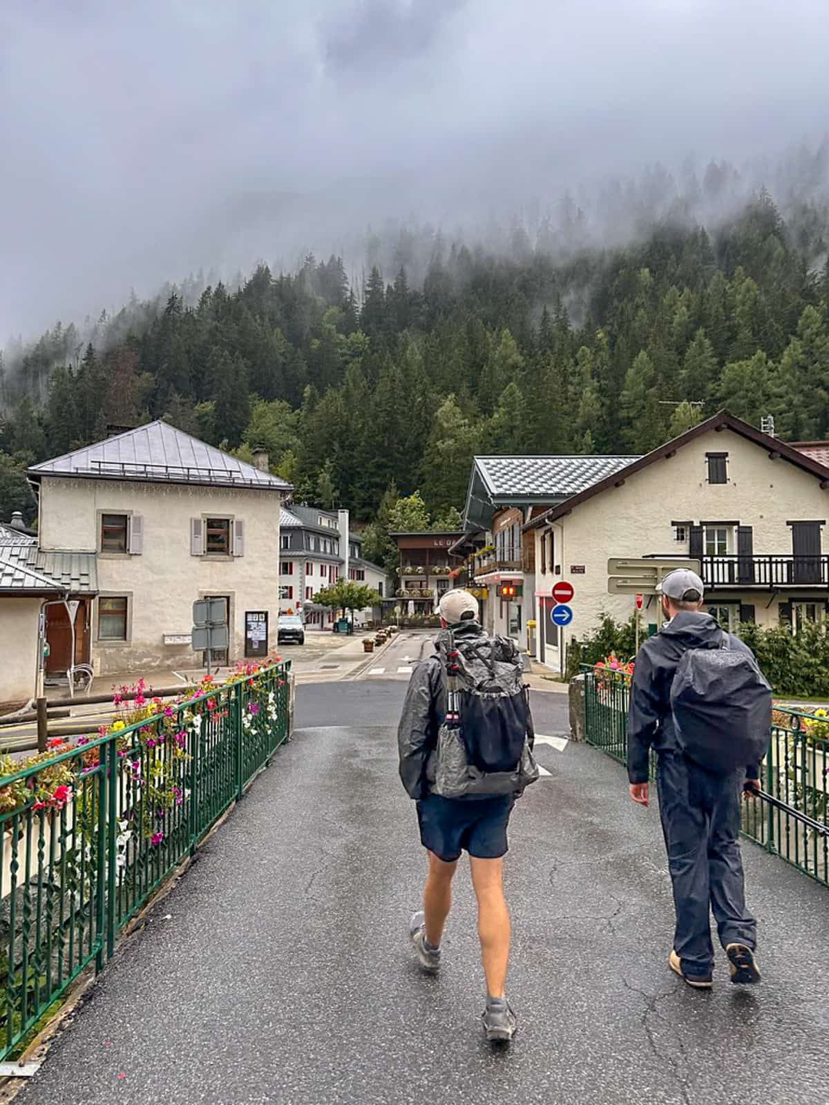 Two men walking along a paved street with French chalets in the background with low-hanging clouds and pine tree forests in the background along the Tour du Mont Blanc