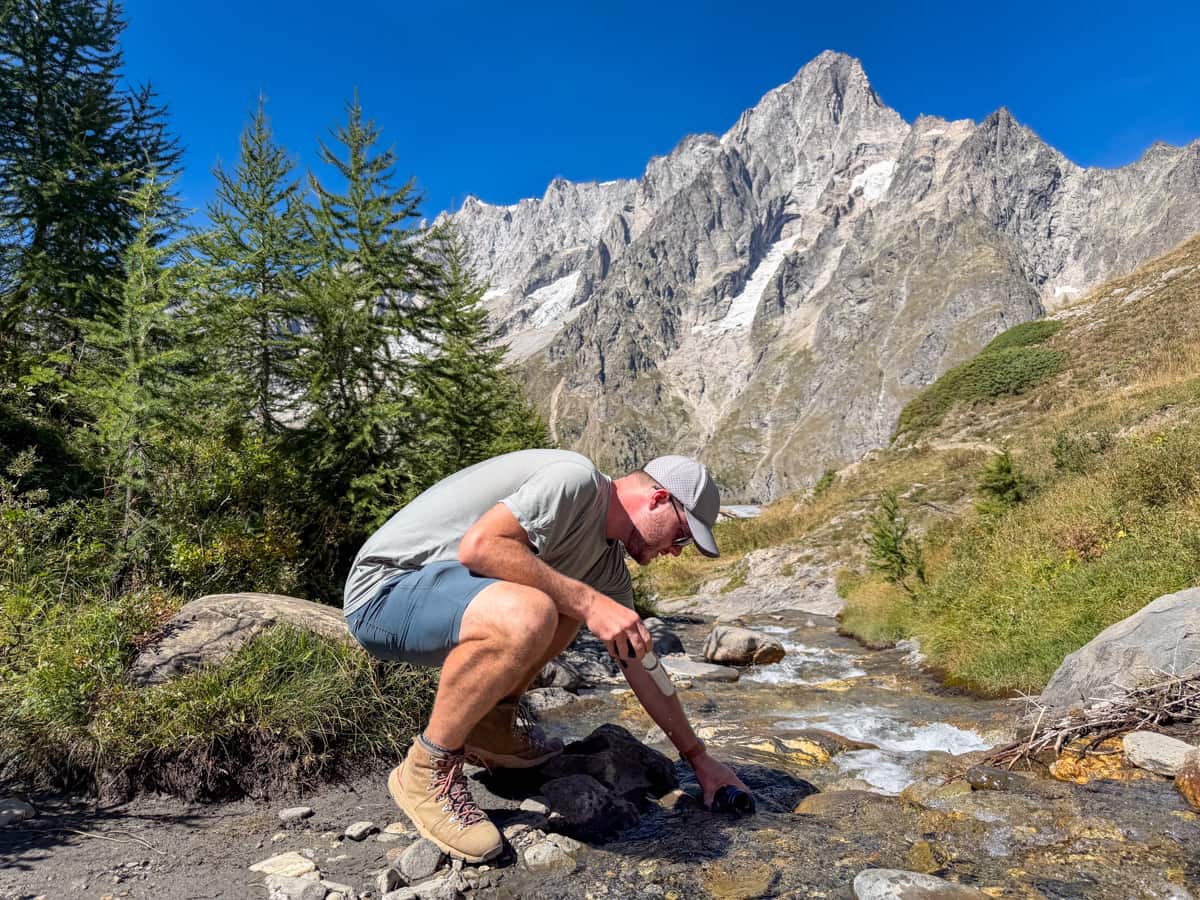 Man filling up a Nalgene bottle in a stream with the Italian Alps in the background near Rifugio Bonatti, Italy along the Tour du Mont Blanc