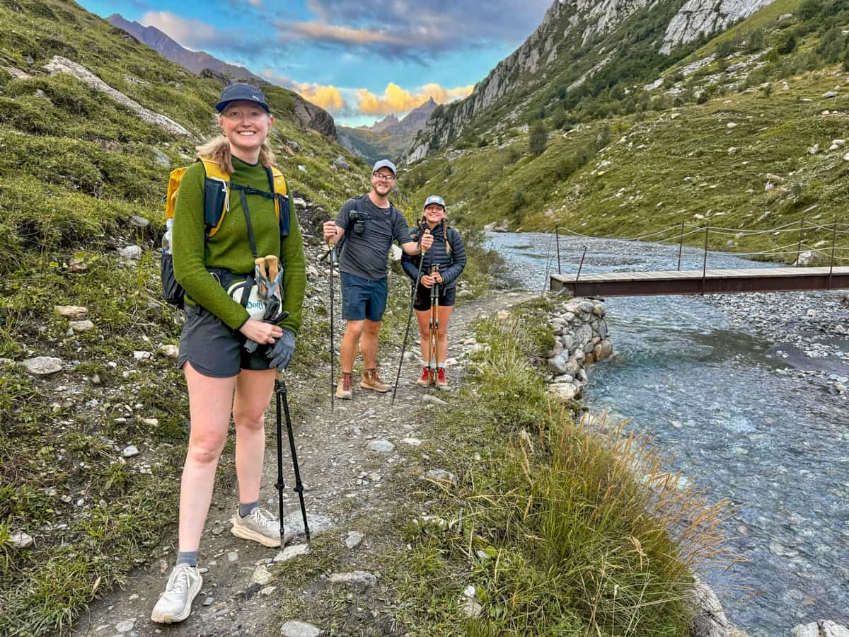 Three hikers smiling on a rocky pathway next to a river near Les Chapieux, France along the Tour du Mont Blanc