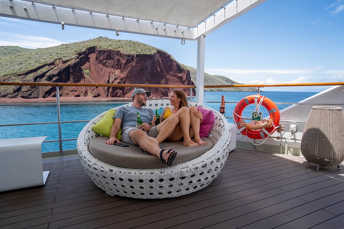 Couple sitting on a lounge chair on top deck of the National Geographic Islander II with Rabida Island in the background on a National Geographic-Lindblad Expeditions cruise in the Galapagos Islands, Ecuador