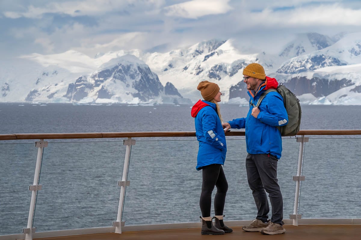 Couple standing on the top deck of the Greg Mortimer ship with snowy mountains in the background on an Antarctica cruise